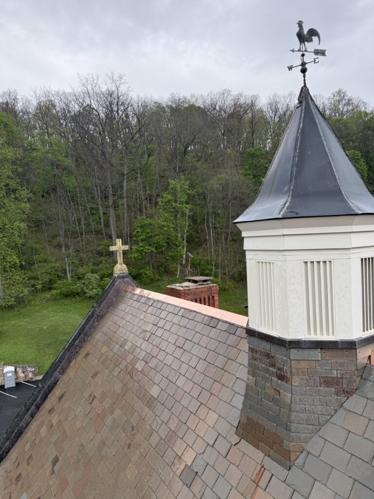 Completed custom copper ridge cap installation on Vermont Gray slate roof at St. Matthew Roman Catholic Church Tyrone PA Blair County showing new copper flashing against historic slate field cross and weather vane by GSM Roofing 2026